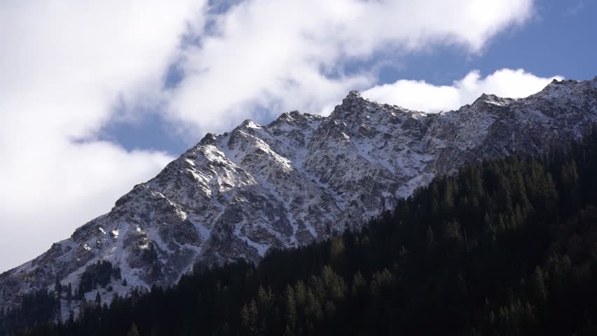 Majestic snow covered mountain peak with a pine forest under a clear blue sky with passing clouds