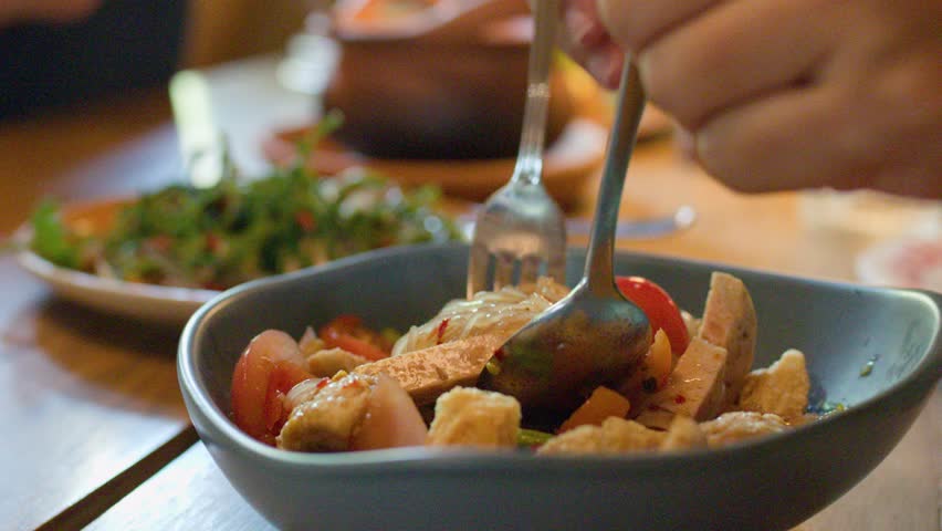 Hands mix spicy noodle salad with tomatoes and pork skin in warmly lit restaurant setting