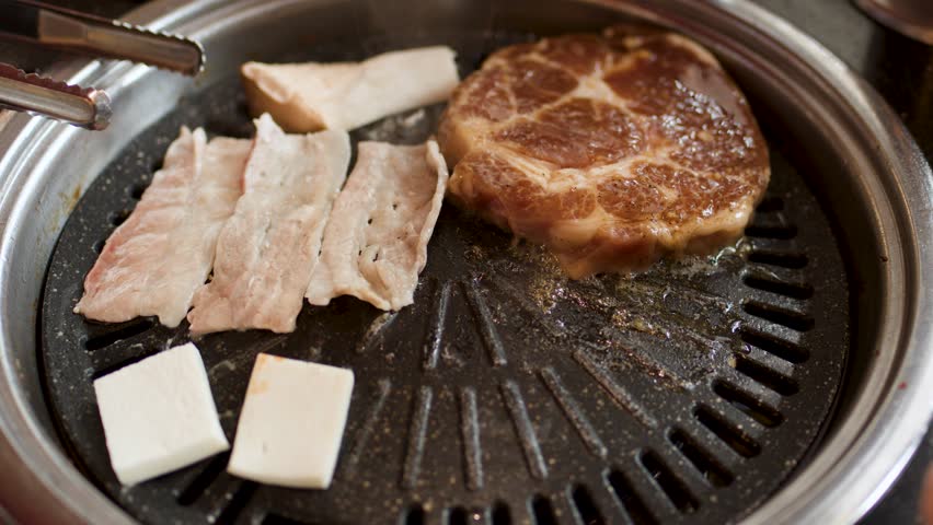 Hand uses tongs to place raw pork belly on hot Korean barbecue grill, natural lighting