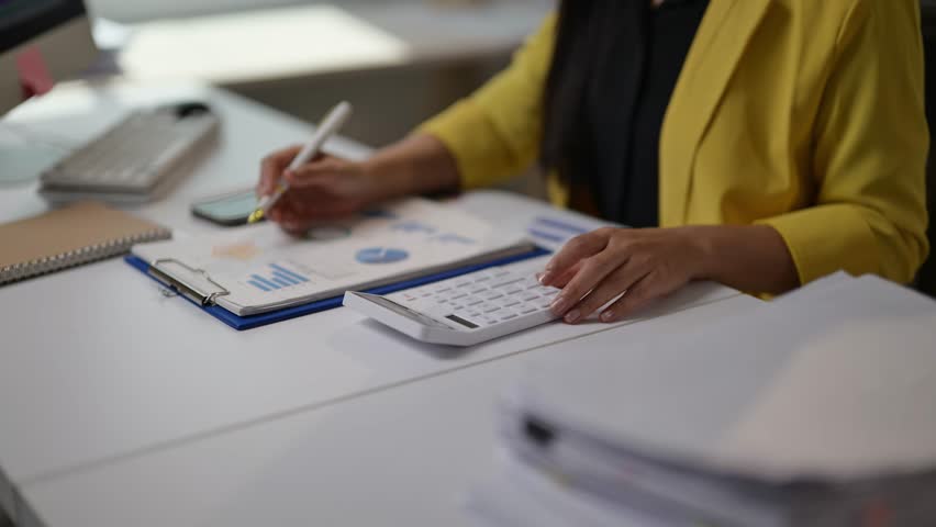 A woman is sitting at a desk with a calculator and a piece of paper with graphs on it. She is writing on the paper with a pen