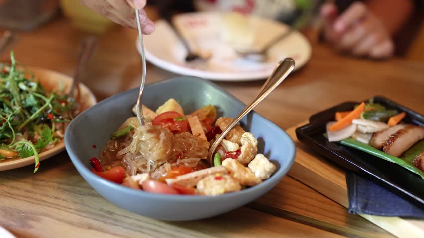 Hand stirs spicy Thai glass noodle salad in bright restaurant, surrounded by fresh side dishes