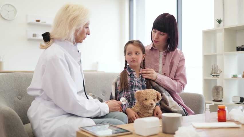 A doctor uses a stethoscope to examine a young girl while her mother comforts her, teddy bear in hand, during a checkup.