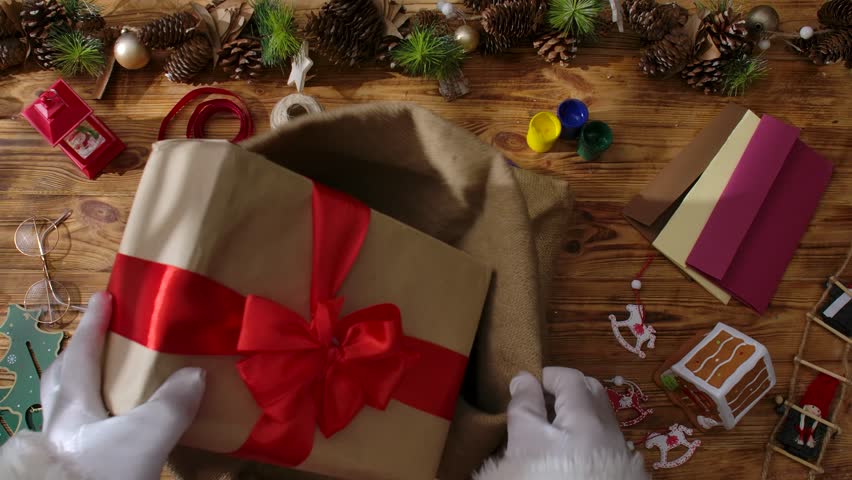 A top view of Santa's gloved hands folding gifts into his sack on a wooden table among cards, paints, and garlands, symbolizing the arrival of Christmas and preparations for the holiday.