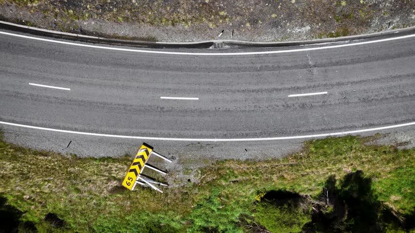Aerial view of a white car driving along a rural road, contrasting with the lush green landscape and a speed limit sign blown over by strong winds, Hanmer Springs, Canterbury Region, New Zealand.