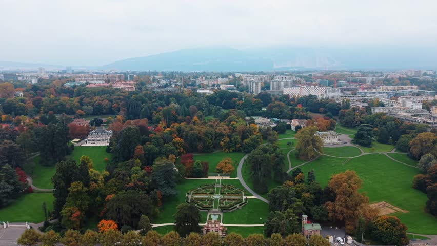 Aerial view of Parc des Eaux-Vives, with its lush green lawns, colorful trees, and ornate gardens, set against the backdrop of Geneva, Switzerland.