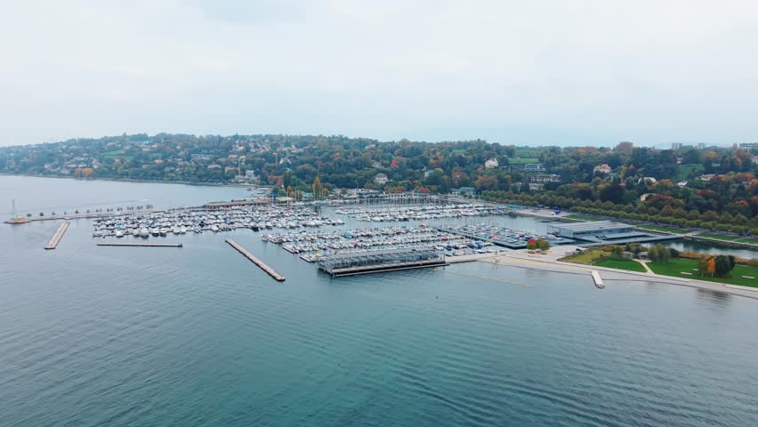 Aerial view of boats docked at Quai Gustave-Ador, where the turquoise water meets the shore and lush trees displaying autumnal colors, Port-Noir, Geneva, Switzerland.