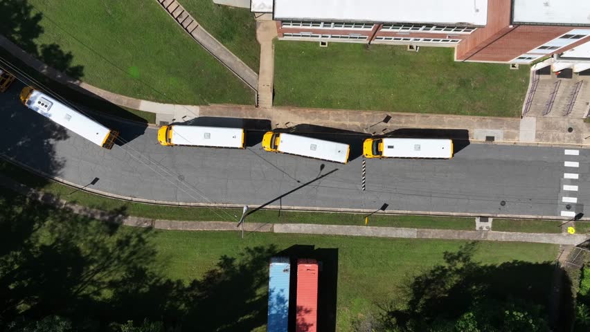 Row of yellow school buses in front of school campus. Arriving at historic school in at midday. Aerial top down shot.