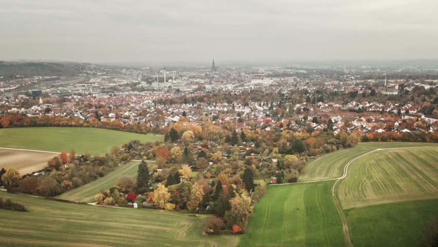 Camera glides forward across green meadows, seasonal gardens, and golden foliage, drawing the eye toward the cityscape of Ulm on the horizon.