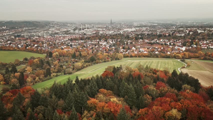 Camera glides right over layers of vibrant autumn foliage, tracing the border between colorful woods, farmlands, and the cityscape of Ulm.