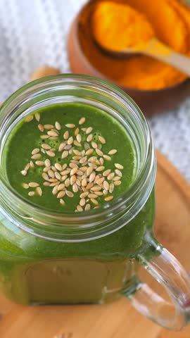 Healthy green smoothie, topped with sesame seeds in a glass jar, rotating on a wooden board with a bowl of turmeric powder in the background, showcasing a nutritious and vegan friendly breakfast