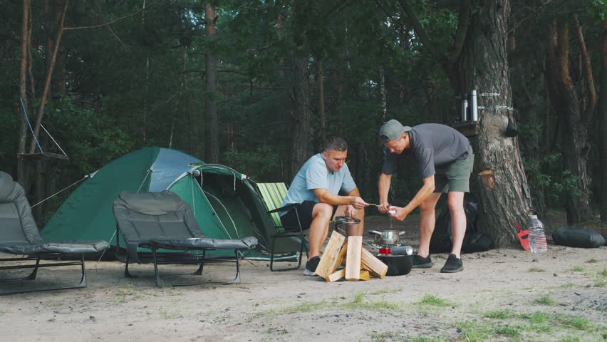 Caucasian friends preparing meal outdoors, Relaxed two men cooking and gathering at riverside campsite, Caucasian males enjoying summer time together building fire on sandy riverbank