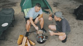 Closeup campfire cooking by tent two caucasian men preparing meal on portable grill arranging wood and utensils near tent entrance sandy ground riverbank backdrop warm summer light focused - Powered by Shutterstock - Get 15% off with code: PIKWIZARD15