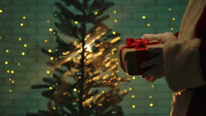 A festive scene featuring a Santa Claus figure holding a wrapped Christmas gift with a red ribbon in front of a beautifully lit Christmas tree.