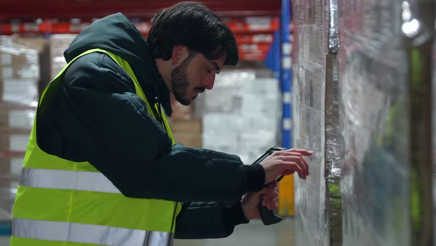 Young warehouse employee in a high visibility vest and warm jacket scanning a pallet of goods wrapped in plastic with a handheld barcode reader in a large, cold distribution center - Powered by Shutterstock - Get 15% off with code: PIKWIZARD15
