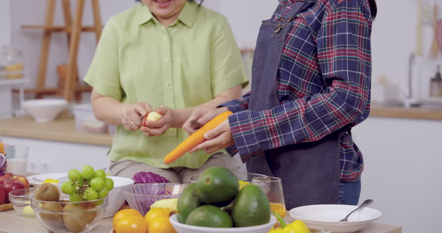 Senior asian mother peel apple while adult daughter hold carrot beside kitchen counter full colorful fruits and vegetables, enjoy family time, teamwork, home cooking promotes health strong bonding