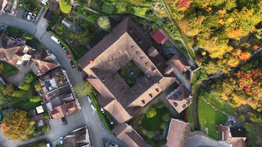 Top-down aerial view of a square, brown-roofed building complex surrounded by a small town and vibrant autumn foliage in Walenstadt, Schweiz.