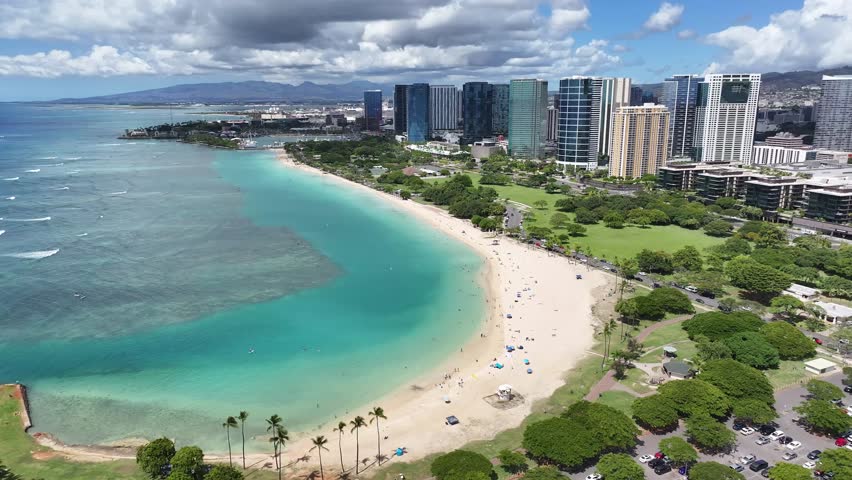 Aerial drone footage of tropical Waikiki Beach in Honolulu, Oahu, Hawaii, showing turquoise ocean waves, palm trees, and hotels along the coastline.