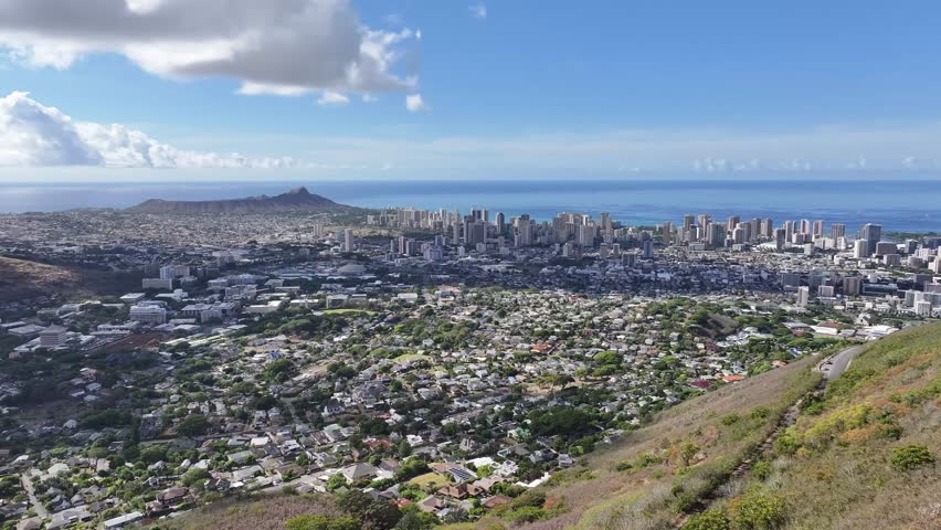 Aerial drone footage overlooking the Honolulu skyline on Oahu, Hawaii, showcasing high-rise hotels, tropical coastline, blue ocean waters, and the scenic cityscape of Waikiki under sunny skies.
