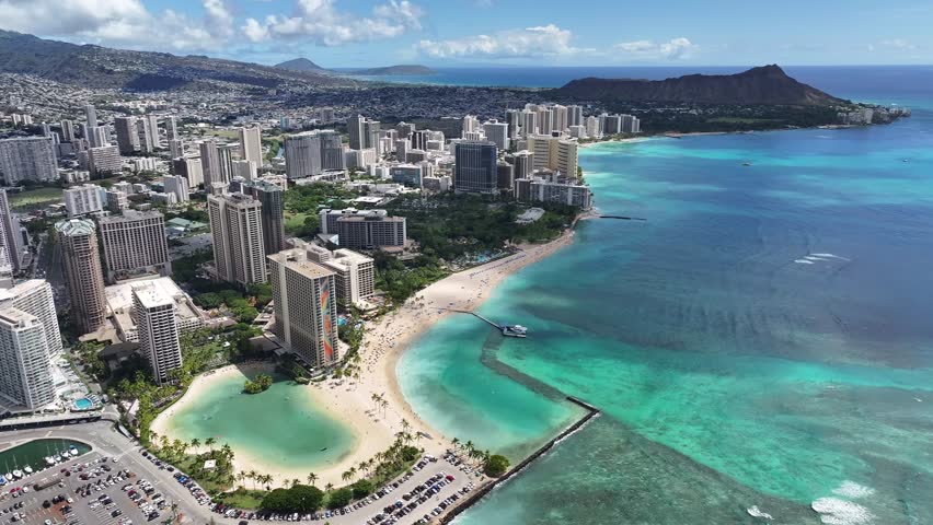 Aerial drone footage over Waikiki Beach in Honolulu, Oahu, Hawaii, showcasing turquoise ocean water, palm trees, hotels, surfers, and the tropical coastline of this world-famous vacation destination.