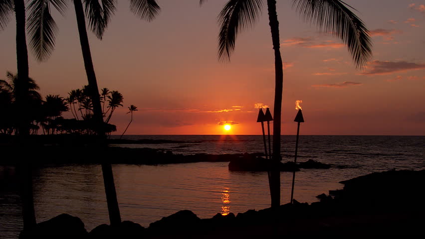 Palm trees and tiki torches sit on the beach at sunset in Hawaii