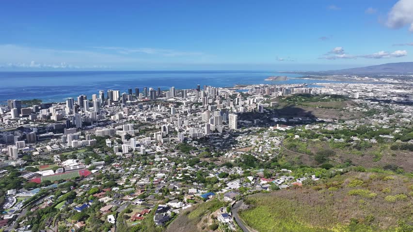 Aerial drone footage overlooking the Honolulu skyline on Oahu, Hawaii, showcasing high-rise hotels, tropical coastline, blue ocean waters, and the scenic cityscape of Waikiki under sunny skies.