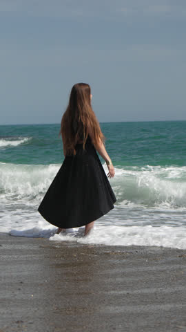 Woman beach ocean, young woman in black dress stands barefoot in ocean waves on sunny day, Vertical video.