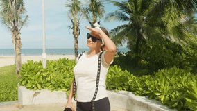 Woman in sunglasses and white headband shades her eyes with a hand while gazing across a sunny tropical beach with palm trees, lush greenery and a relaxed vacation vibe - Powered by Shutterstock - Get 15% off with code: PIKWIZARD15