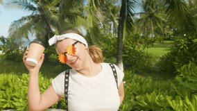 Young woman wearing unique star shaped sunglasses and a bandana, holding a coffee cup, dancing happily and celebrating summer vacation on a tropical beach near palm trees and ocean - Powered by Shutterstock - Get 15% off with code: PIKWIZARD15