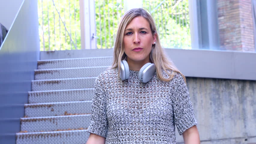 Serious caucasian woman stands on outdoor concrete steps wearing headphones and casual clothes, extending her arm with a thumbs down gesture, expressing disapproval or refusa.