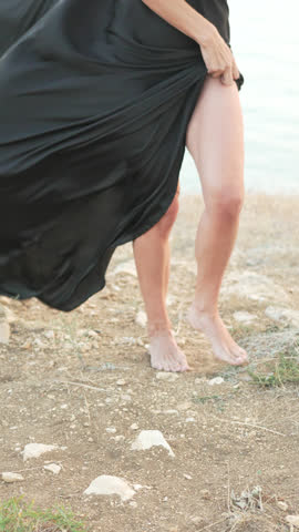 Woman dress sea, elegant barefoot woman in black dress on rocky shore, wind blowing, serene mood. Vertical video.