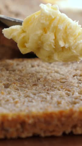 Delicious yellow butter being spread with a knife on a slice of fresh whole wheat bread, creating a simple and wholesome snack or breakfast meal with creamy dairy and healthy whole grains