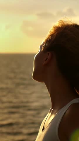 Serene woman with windswept hair looking at the golden sunset reflecting on the calm sea, embracing a moment of peace, tranquility, and contemplation during a beautiful summer evening