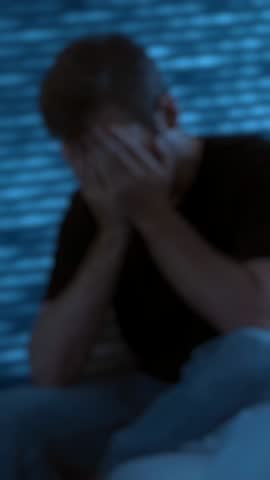 Blurred image, a young man sits on the edge of his bed in a dark room at night, holding his head in his hands, overwhelmed by stress, anxiety, and mental health struggles. vertical video