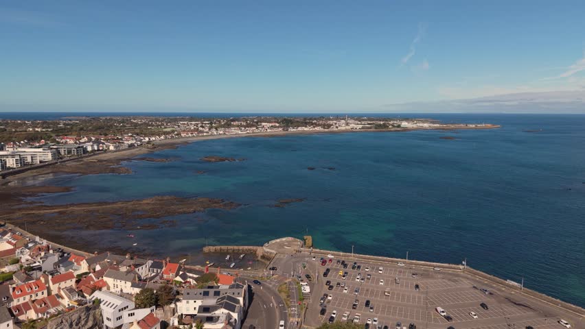 High drone footage over Belle Greve Bay Guernsey at high tide flying out over Salerie Corner and showing rocks,beach ,clear blue water and distant views of St Sampson on sunny day