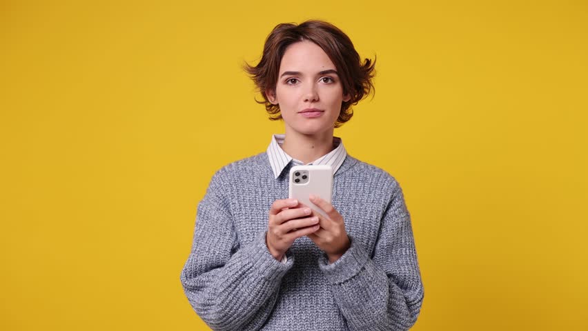 Happy calm young woman wear grey knitted sweater white shirt casual clothes hold use mobile cell phone typing browsing chatting send sms isolated on plain yellow background studio. Lifestyle concept