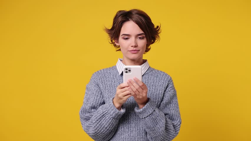 Happy excited surprised young woman wear grey knitted sweater white shirt casual clothes hold use point finger mobile cell phone just found out great big win news isolated on plain yellow background