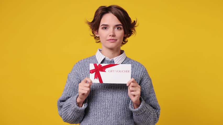 Young woman wear grey knitted sweater white shirt casual clothes pointing finger on gift certificate coupon voucher card for store show thumb up isolated on plain yellow background. Lifestyle concept