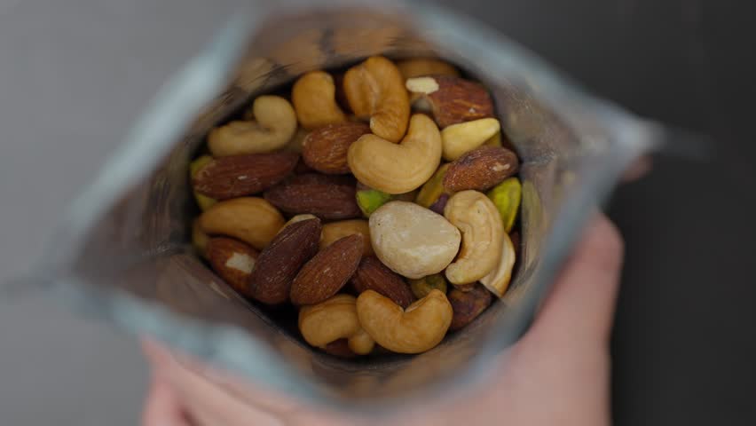 A person hand is picking up a bag of roasted nuts, almonds, cashews, macadamia nuts, and pistachios to eat