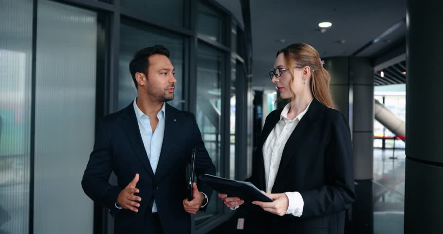 Confident experienced boss explains project details and gives constructive feedback to focused female employee while walking together in modern office corridor