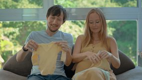 Smiling couple expecting a baby sit on a couch holding baby clothes, enjoying a tender moment as they prepare to welcome their newborn. Slowmotion video. - Powered by Shutterstock - Get 15% off with code: PIKWIZARD15