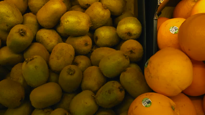 Dolly top down view glides smoothly over piles of fresh oranges, kiwis and lemons stacked in separate sections of market boxes. Fruits are layered on top of each other, textures and colors in close up