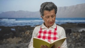 Senior woman with grey hair wearing apron reading a book at seaside beach with mountains in background. - Powered by Shutterstock - Get 15% off with code: PIKWIZARD15