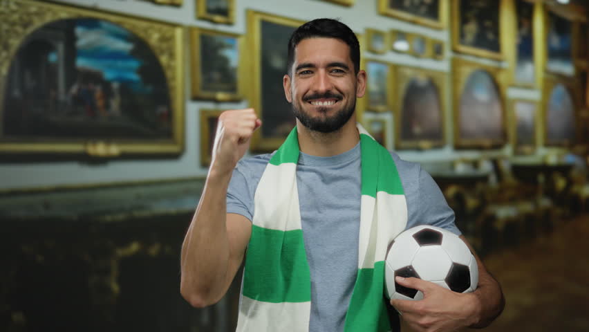 Young man with a soccer ball and scarf smiles confidently inside a museum filled with framed paintings, representing enthusiasm and style in an artistic environment.