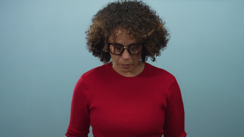 Hispanic woman with curly hair wearing glasses holds red heart close to face with a smile in blue studio; love joy.