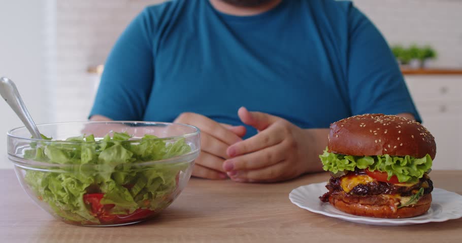 Overweight hungry man choosing diet, healthy salad or unhealthy burger choice. Hands of person with nutrition dilemma making pause above bowl to compare options, guy with big tummy sitting at table
