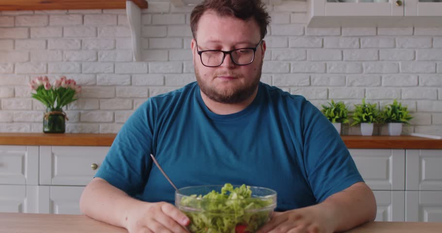 Obese man eating salad in home kitchen, thinking about healthy vegetarian diet and food balance. Plus size adult person with glasses and beard choosing to lose weight and bowl of green lettuce