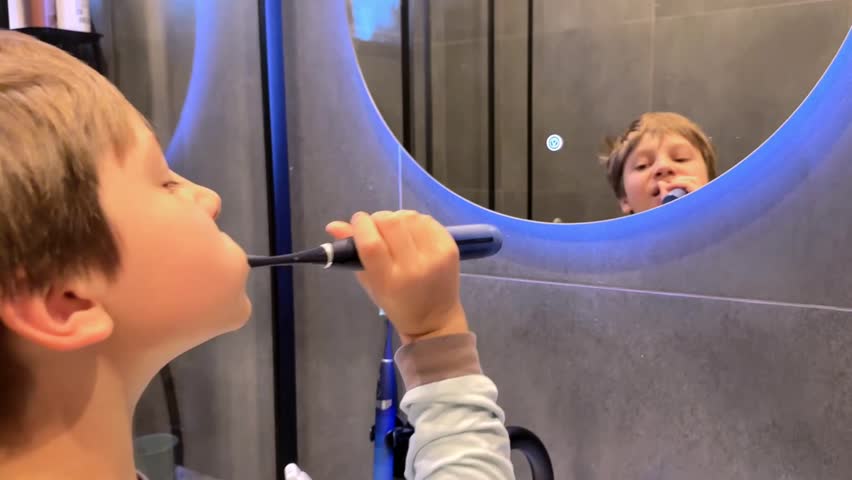 8 year old boy in modern bathroom brushing teeth with electric brush. face reflection in a mirror