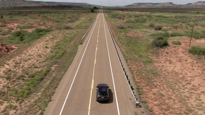An aerial shot of a car driving on a desert highway in New Mexico, USA, showcasing vast landscapes, mesas, and a long, empty road under a clear sky.
