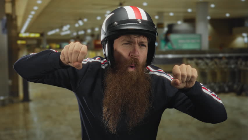 Bearded man wearing red and white helmet grips invisible handlebars and mimics motorcycle riding in an airport terminal; playfulness.