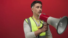 Young man wearing a reflective vest passionately uses a megaphone against a striking red wall, embodying the spirit of volunteerism and communication. - Powered by Shutterstock - Get 15% off with code: PIKWIZARD15